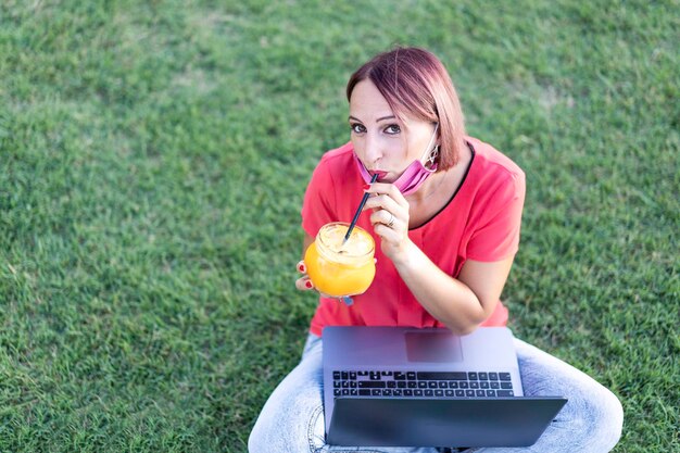 Woman working outdoors with laptop during coronavirus outbreak Smiling female entrepreneur sitting on lawn with pc while drinking health orange juice for break Remote work concept into the nature