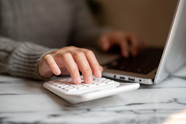 A woman working at a table indoors typing on a laptop keyboard and using a calculator