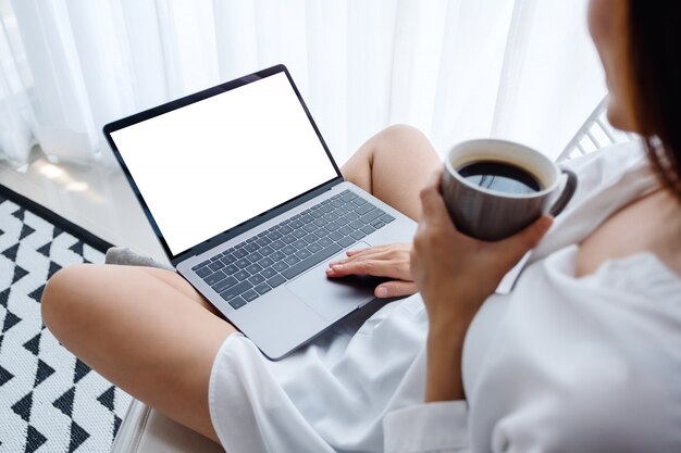  of a woman working and typing on laptop computer with blank screen while sitting and drinking coffee in a bedroom at home