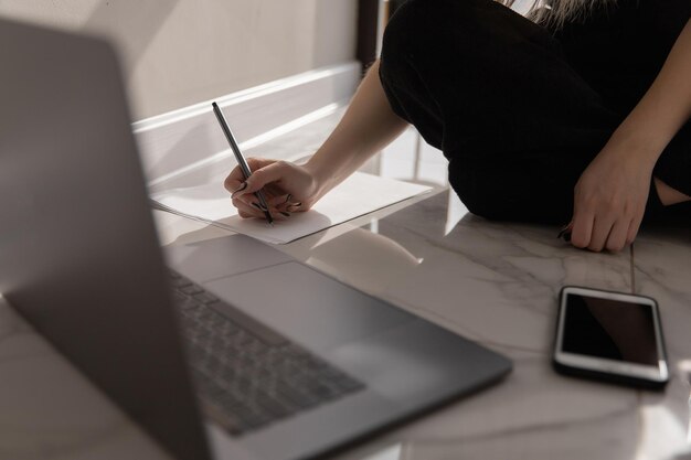 A woman works at home at a computer sitting on the floor by the window and fills out paperwork