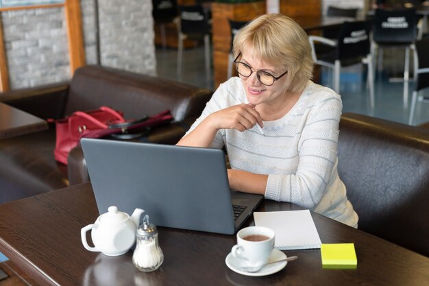 A woman works at a laptop in a cafe. Middle-aged woman drinking tea and watching the news. She's a freelancer.