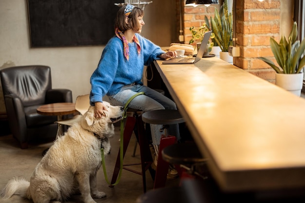 女性は、モダンなコーヒー ショップで犬と一緒に座ってノート パソコンで作業します。