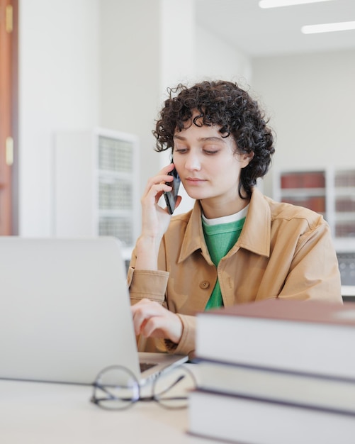 Woman works in library coworking at laptop and talks on phone researcher at university