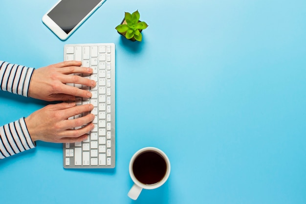 Woman works in the office on a blue background.Only hands Concept workspace, working at a computer, freelance, design. phone, flower, pencil. Banner. Flat lay, top view