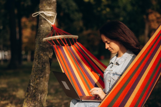 A woman works remotely from a laptop while lying in a hammock in the park
