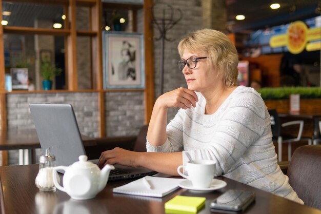 A woman works with a laptop at a table in a cafe or in the office. A middle-aged woman works remotely. She's a businesswoman.