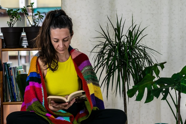 Foto una donna avvolta in una coperta colorata con i piedi nudi siede a leggere un libro nella sua confortevole casa