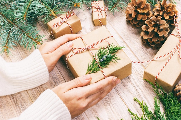 크리스마스 선물을 포장하는 여자, 전나무와 소나무 콘으로 크리스마스 선물을 준비합니다. 크리스마스 장식으로 나무 배경에 손으로 만든 선물. 상위 뷰, 복사 공간입니다. 톤.