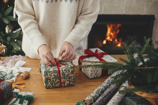 Woman wrapping stylish christmas gift with red ribbon on wooden table with festive decorations against fireplace in decorated scandinavian room Merry Christmas Hand holding present