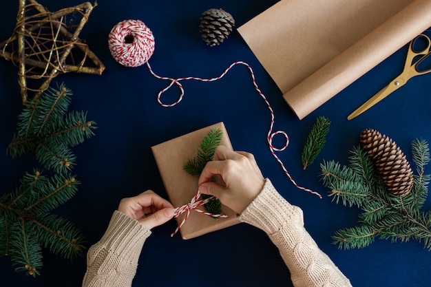 写真 女性は青い木製の背景にクラフト紙と天然トウヒの枝で手作りのエコスタイルのクリスマスプレゼントを包みます。