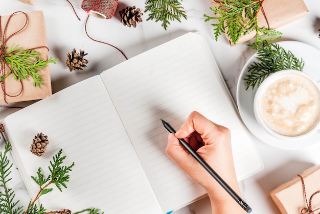 Woman write wishes or to do list in notebook, coffee mug,  gift or present box, decorated with christmas tree branches, pine cones, red berries, on white marble table,  top view