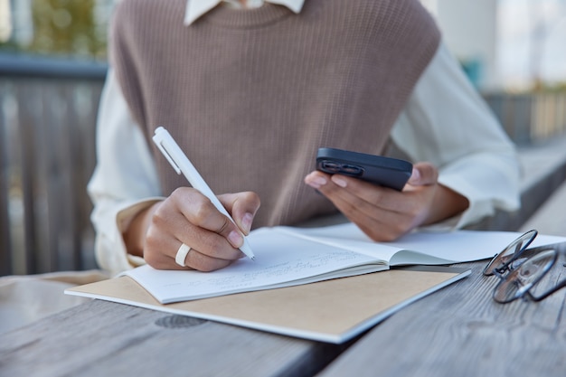 Женщина делает заметки тезис для исследовательского отчета держит эссе современный мобильный телефон держит ручку составляет список или план занимается тайм-менеджментом сидит за деревянным столом на открытом воздухе