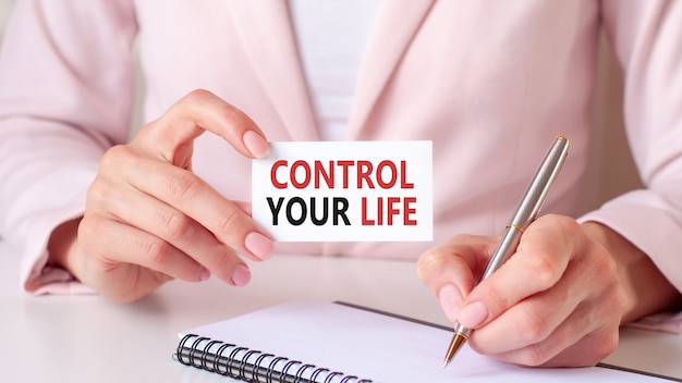 Женщина пишет в блокноте серебряной ручкой и рукой держит карточку с текстом: контролируйте свою жизнь