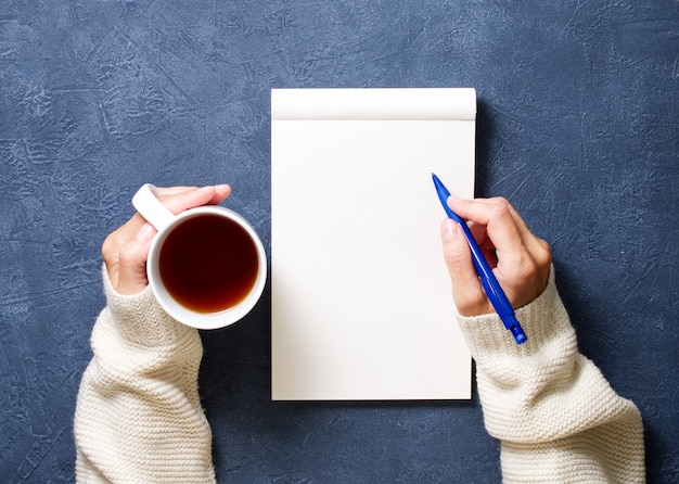 Woman writes in notebook on dark blue table, hand in shirt holding a pencil, cup of tea, 