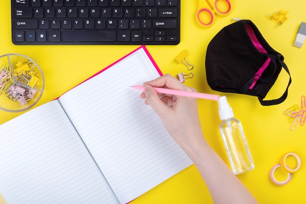 Woman writes in notebook at desk, next to antiseptic and mask, yellow background.