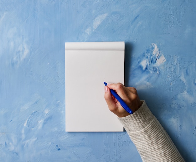 Woman writes in notebook on stone blue table, hand in shirt holding a pencil,