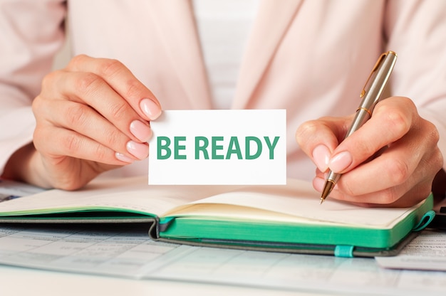 Woman writes in a notebook with a silver pen and hand hold card with text: BE READY