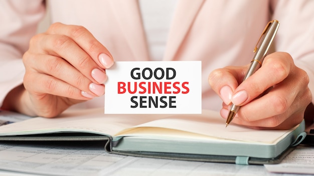 Woman writes in a notebook with a silver pen and hand hold card with text: good business sense. Pink background, front view. Business and education concept.