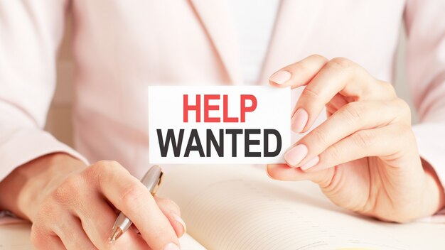 Woman writes in a notebook with a silver pen and hand hold card with text: HELP WANTED