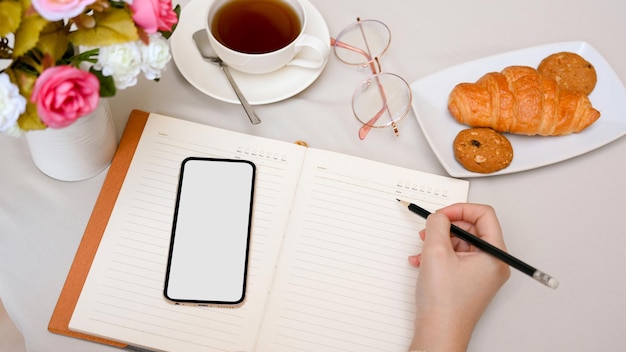 A woman writes something in her diary book with a pencil and a smartphone on the coffee table