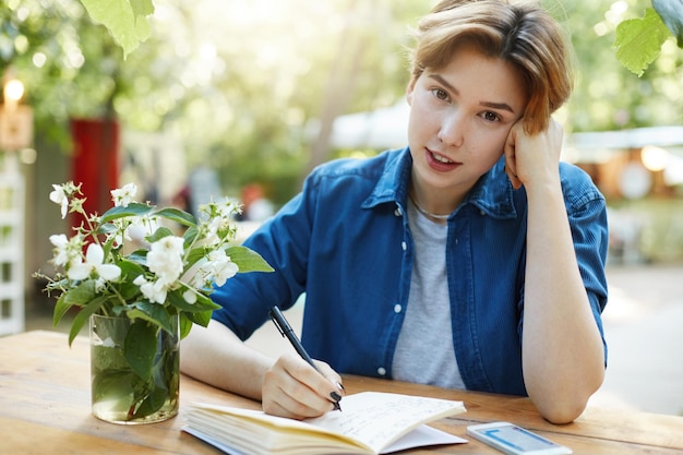 Woman writing in diary Portrait of a girl taking notes in her notepad outdoors planning a trip or vacation or future business looking at camera wearing velvet blue shirt in park