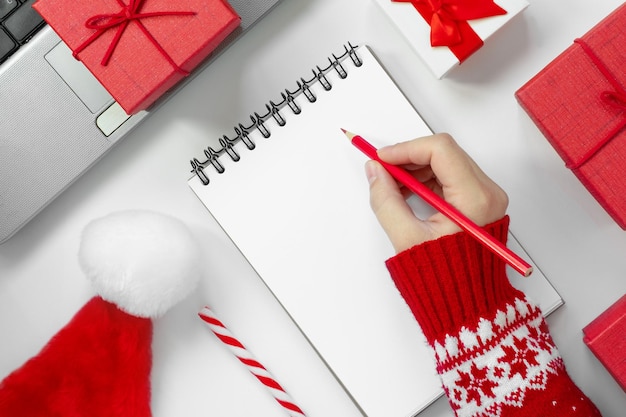 Woman Writing in an Empty Notebook on White Desk Girl's Hands with Pencil Among Red Gift Box and Winter Festive Decor Concept of Diary Holiday Greetings or Planning Top view Mock up Paper Blank