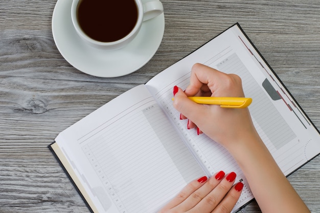 Woman writing essay in notepad at the table, cup of coffee are on the background, overhead view