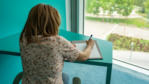 Женщина пишет за современным столом рядом с большим окном с видом на улицу