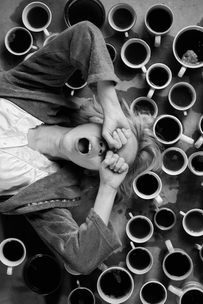 Женщина зевает в окружении множества чашек кофе Крайний срок сверхурочной работы Желание поспать после кофе Нарушение циркадного ритма