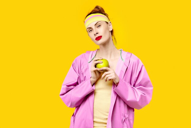 Woman on yellow background and a pink coat is holding an apple