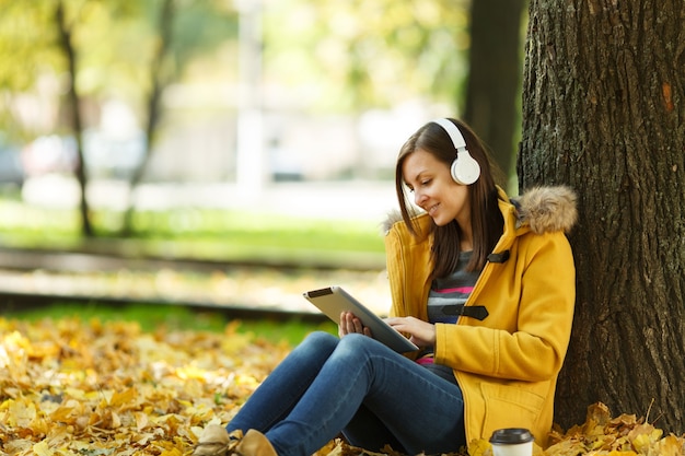 暖かい日に秋の都市公園で、コーヒーやお茶を片手に座って、タブレットを手に、ヘッドフォンを持って木の下で音楽を聴いている黄色いコートとジーンズの女性。秋の黄金の葉。