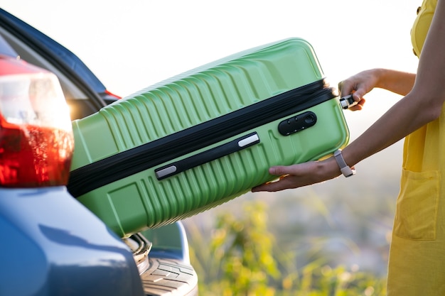 A woman in yellow dress taking green suitcase from car trunk. Travel and vacations concept.