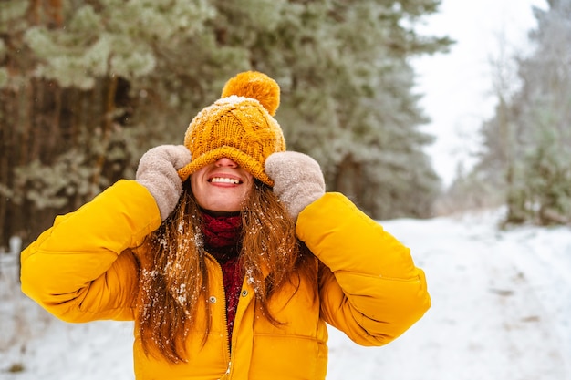 woman in yellow hat is fooling around and pulling a winter hat over her face in winter forest