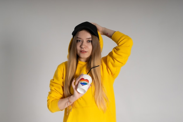 A woman in a yellow hoodie, a black cap with a pierced nose, holds a paper cup with the inscription love is love and a rainbow lgbtq in the shape of a heart