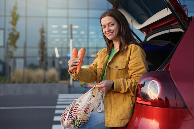 Woman in yellow jacket hold carrots and shopping bag with fruits sitting on red cars trunk