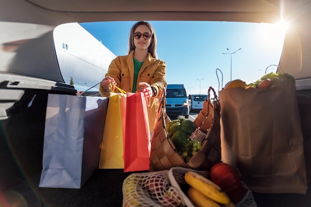 Woman in yellow jacket put shopping bags with a groceries in car trunk