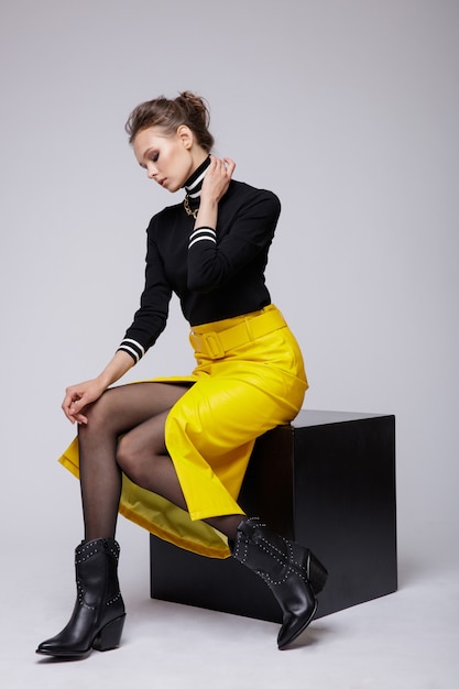 白い背景の上の黄色い革のスカートの黒いブラウスの女性スタジオショットキューブに座って