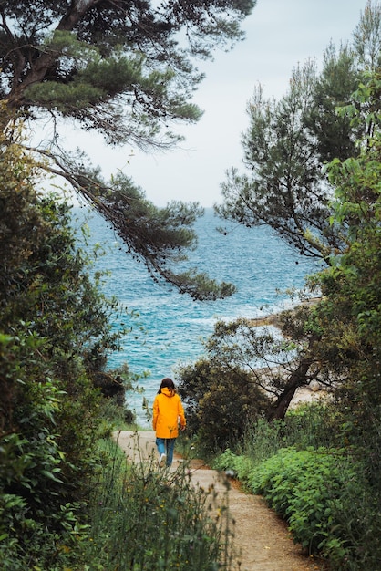 Foto donna in impermeabile giallo che cammina verso il mare da un sentiero tra gli alberi