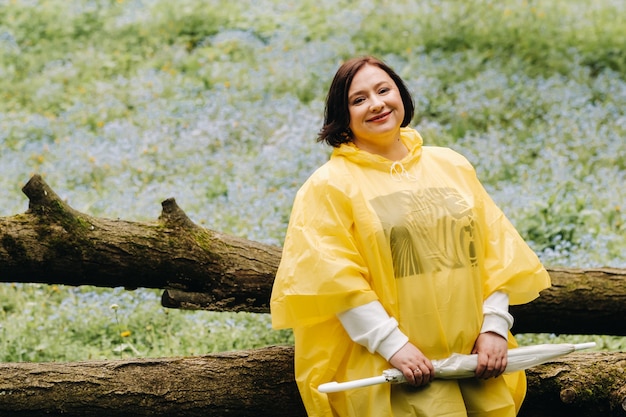 우산을 쓴 노란 우비를 입은 여자가 여름 숲 속 쓰러진 나무에 앉는다.