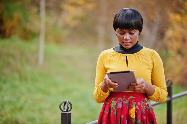 Woman in yellow and red dress at golden autumn fall park with tablet at hands