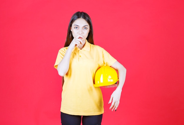 Donna in camicia gialla che tiene un casco giallo e sembra confusa e pensierosa.