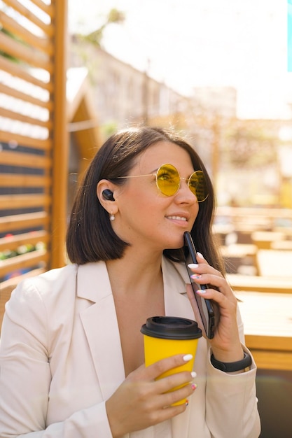 Woman in yellow sunglasses with earbuds in ears with phone glass of coffee looks up smiling