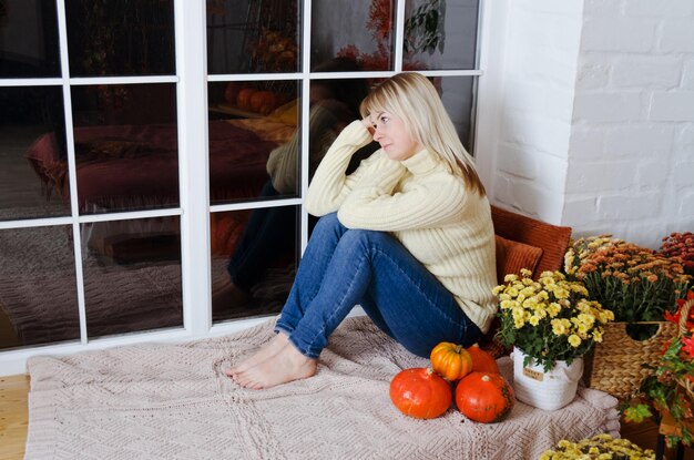 woman in yellow sweater sits on window seat in cozy home with autumn decorations, orange pumpkins