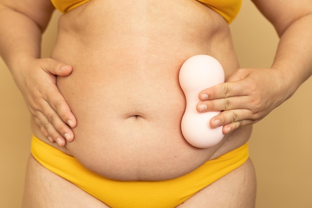 Woman in yellow swimsuit rub thick sagging stomach with body massage roll closeup beige background Slimming fighting overweight obesity and cellulite Plus size people and body positive concept