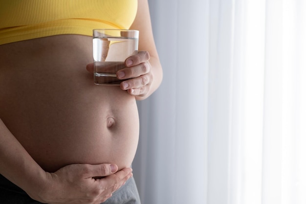 Woman in yellow top with pregnant belly hold glass of water in hands Baby expectation water drinking care and wellness on pregnancy time