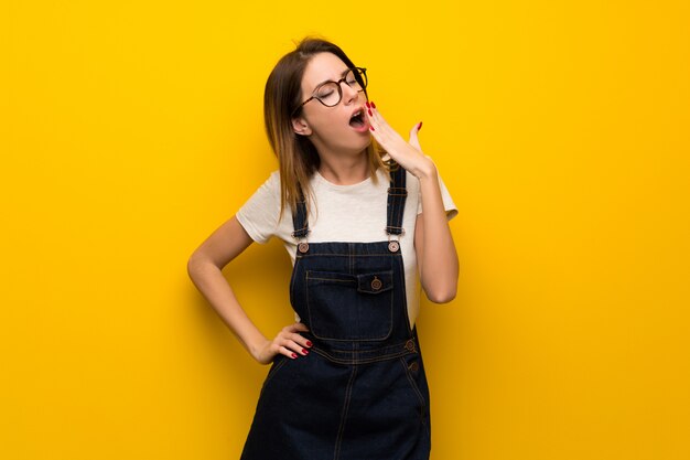 Woman over yellow wall yawning and covering wide open mouth with hand