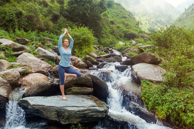 Woman in yoga asana Vrikshasana tree pose at waterfall outdoors