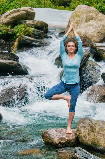 Woman in yoga asana Vrikshasana tree pose at waterfall outdoors