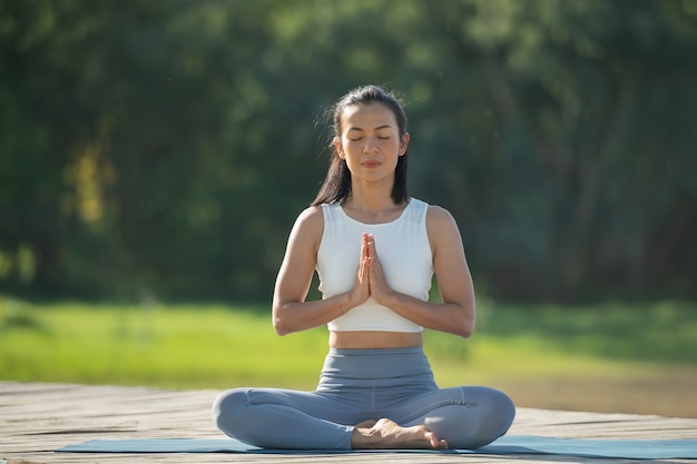 Woman on a yoga mat to relax in the park at mountain lake. Calm woman with closed eyes practicing yoga, sitting in Padmasana pose on mat, Lotus exercise, attractive sporty girl in sportswear.