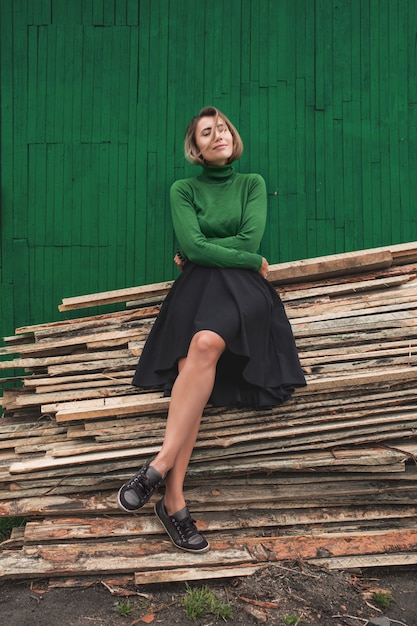 Woman young adult beautiful in a green jumper against a green fence sitting on wooden boards